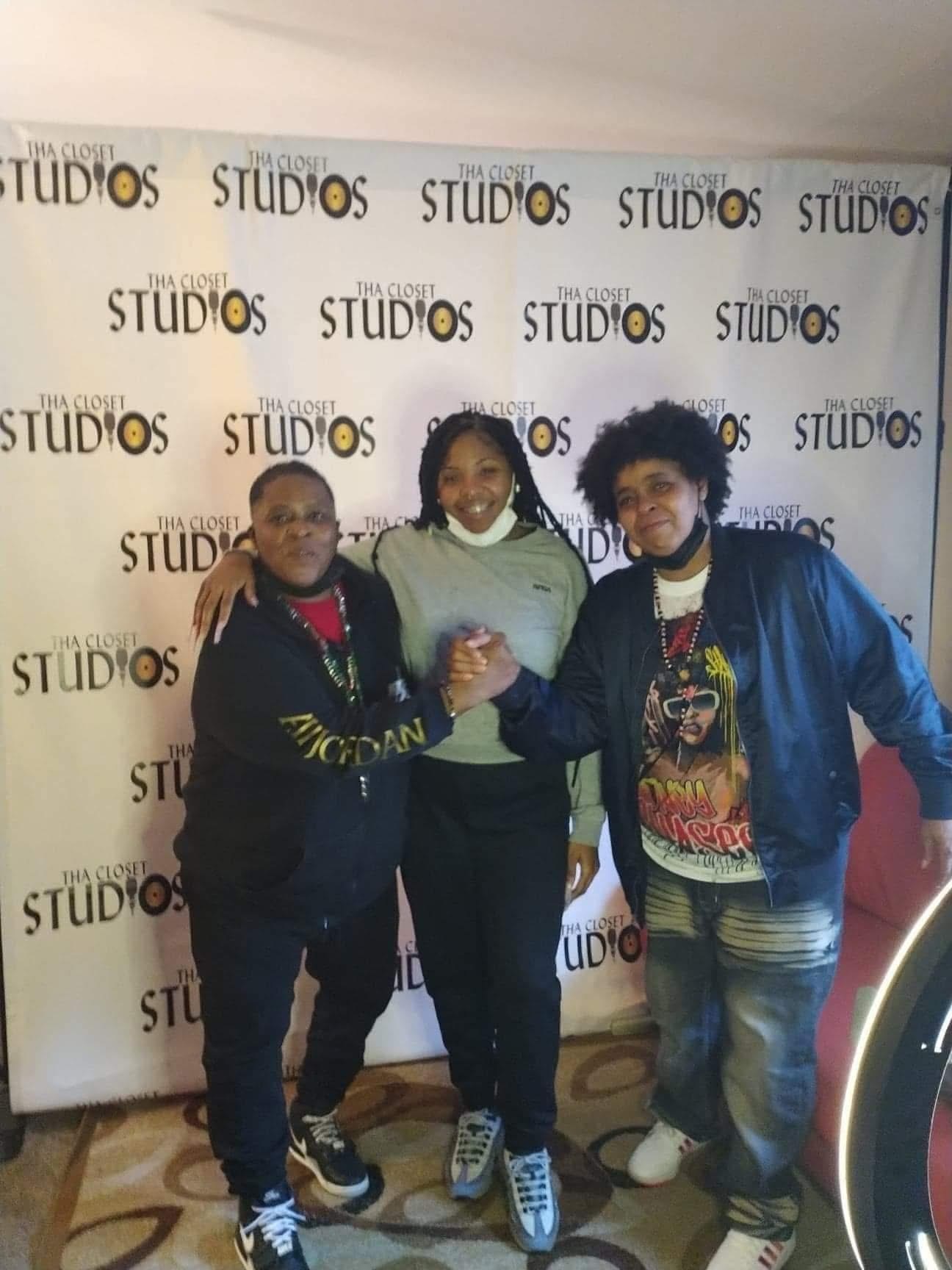 Three people standing together in front of a backdrop with 'STUDIOS' logo, smiling and posing for the photo.