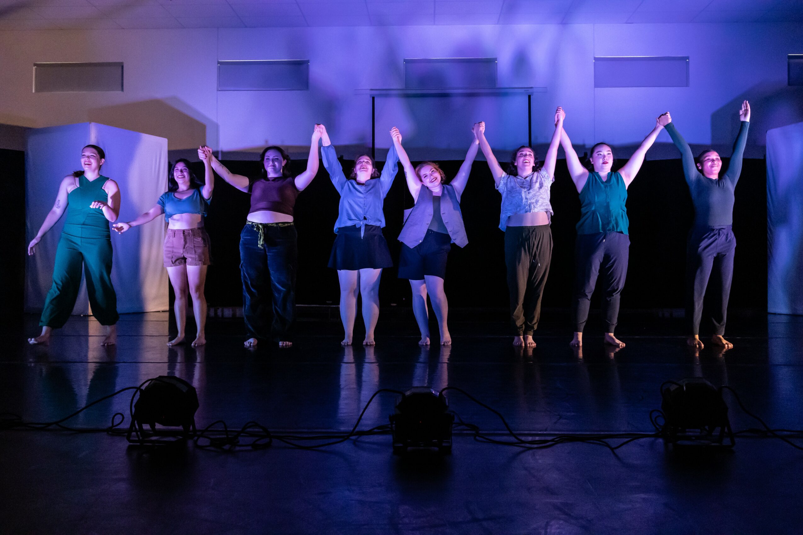 Group of nine dancers holding hands on stage, illuminated by purple lighting, with a dark background and stage equipment.