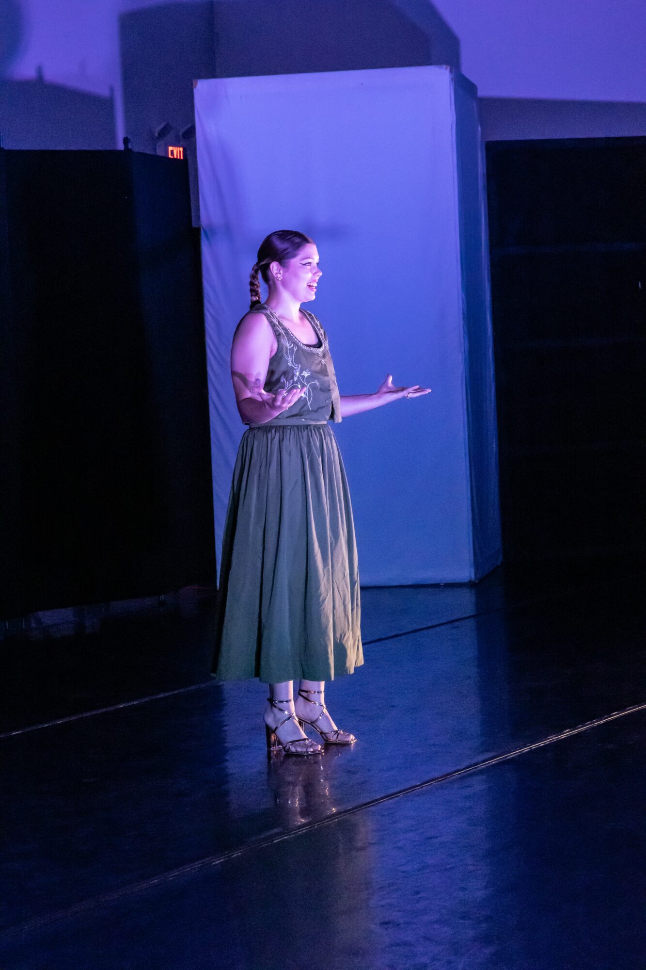Woman standing on stage, speaking or performing, with a blue backdrop and dark surroundings.