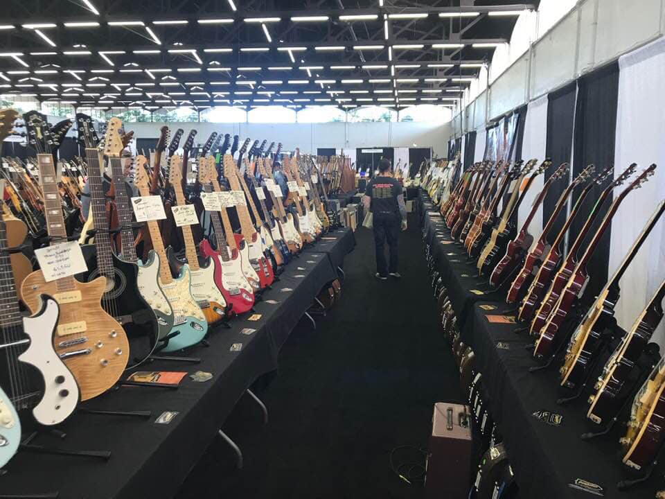 Guitars displayed on tables in a large indoor exhibition space with people browsing.