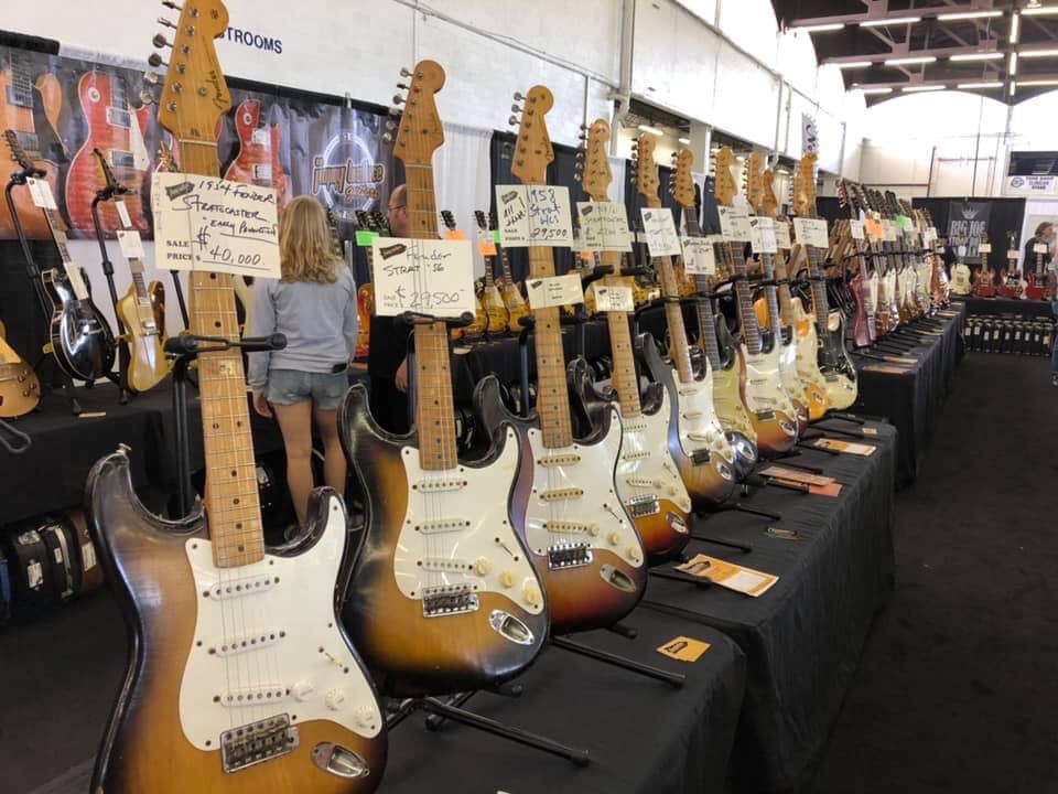 Multiple electric guitars displayed on a table at an exhibition, with a person browsing in the background.