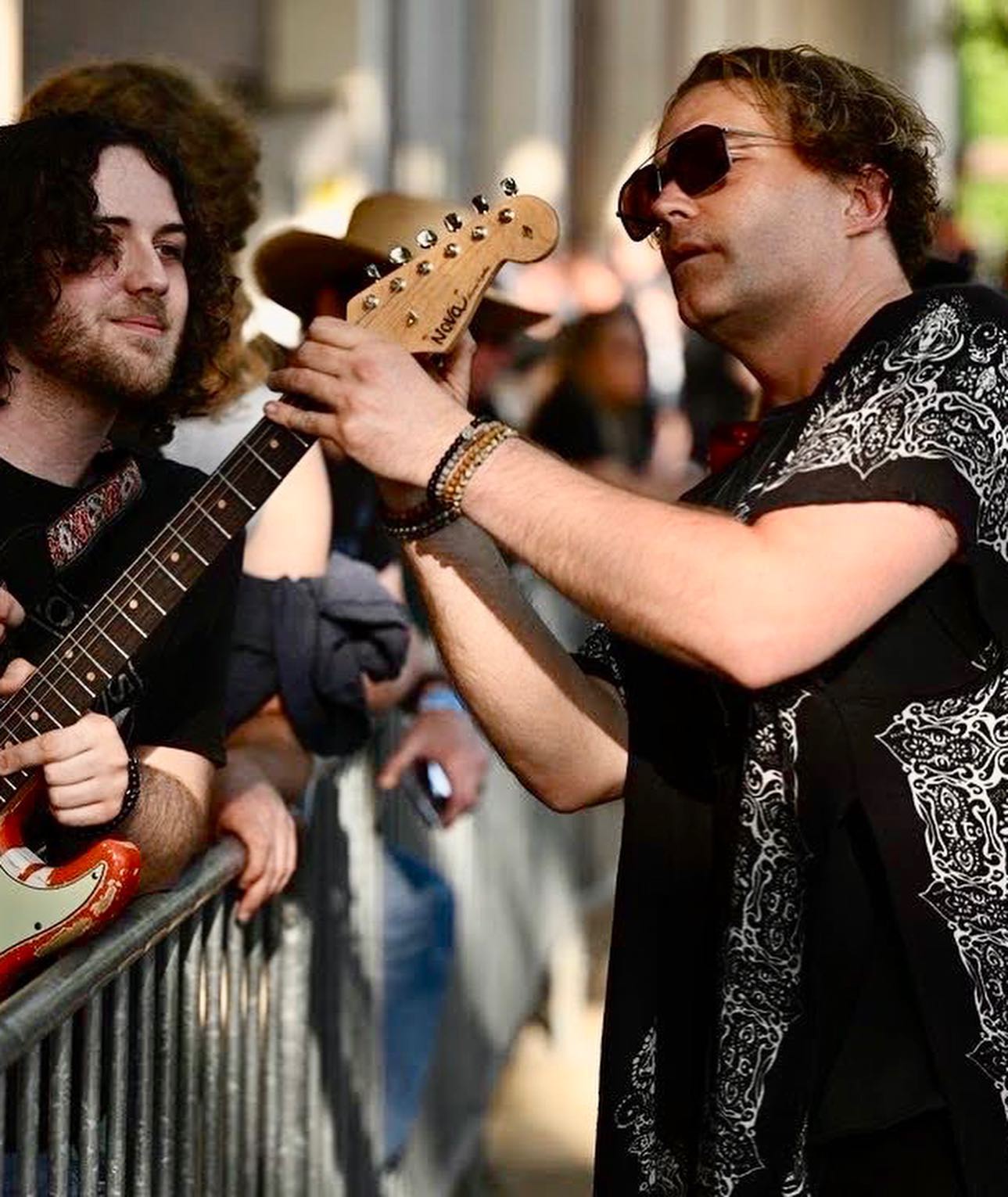 Two men with guitars, one wearing sunglasses, in a crowded outdoor setting.