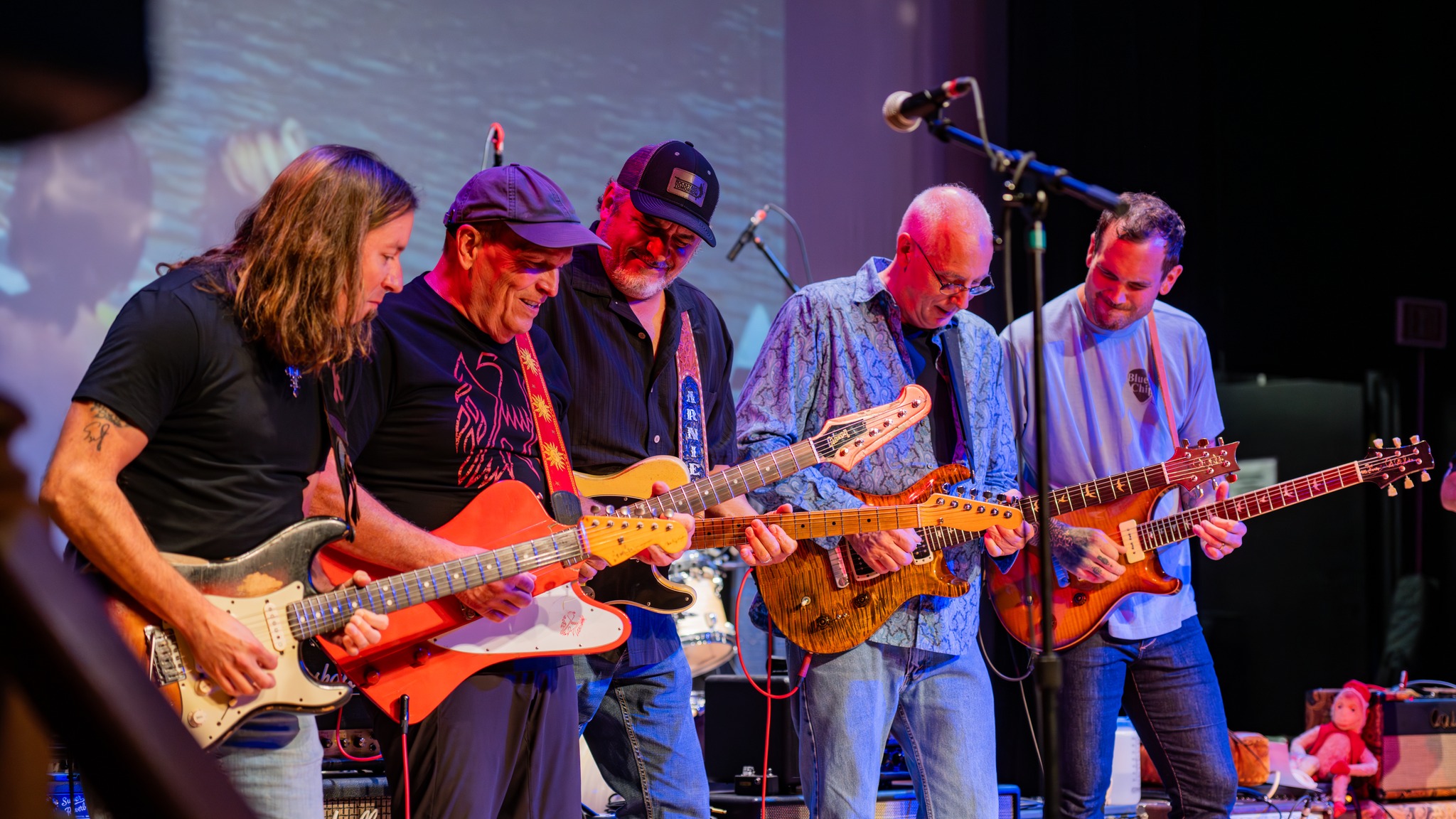 Five musicians playing electric guitars on stage with microphones and colorful background lighting.