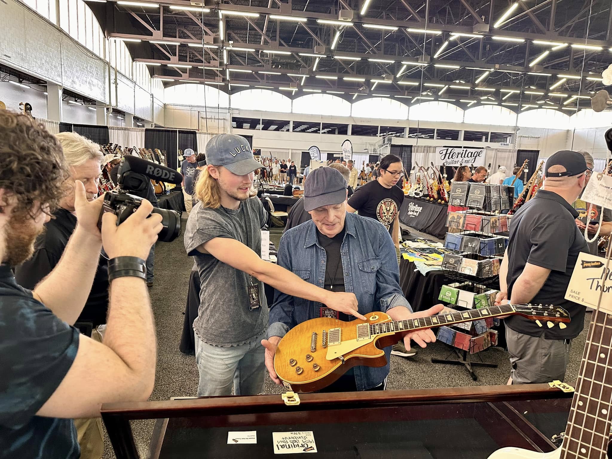 Person with camera filming two people, one holding a yellow guitar, at an indoor market or event with booths and people.