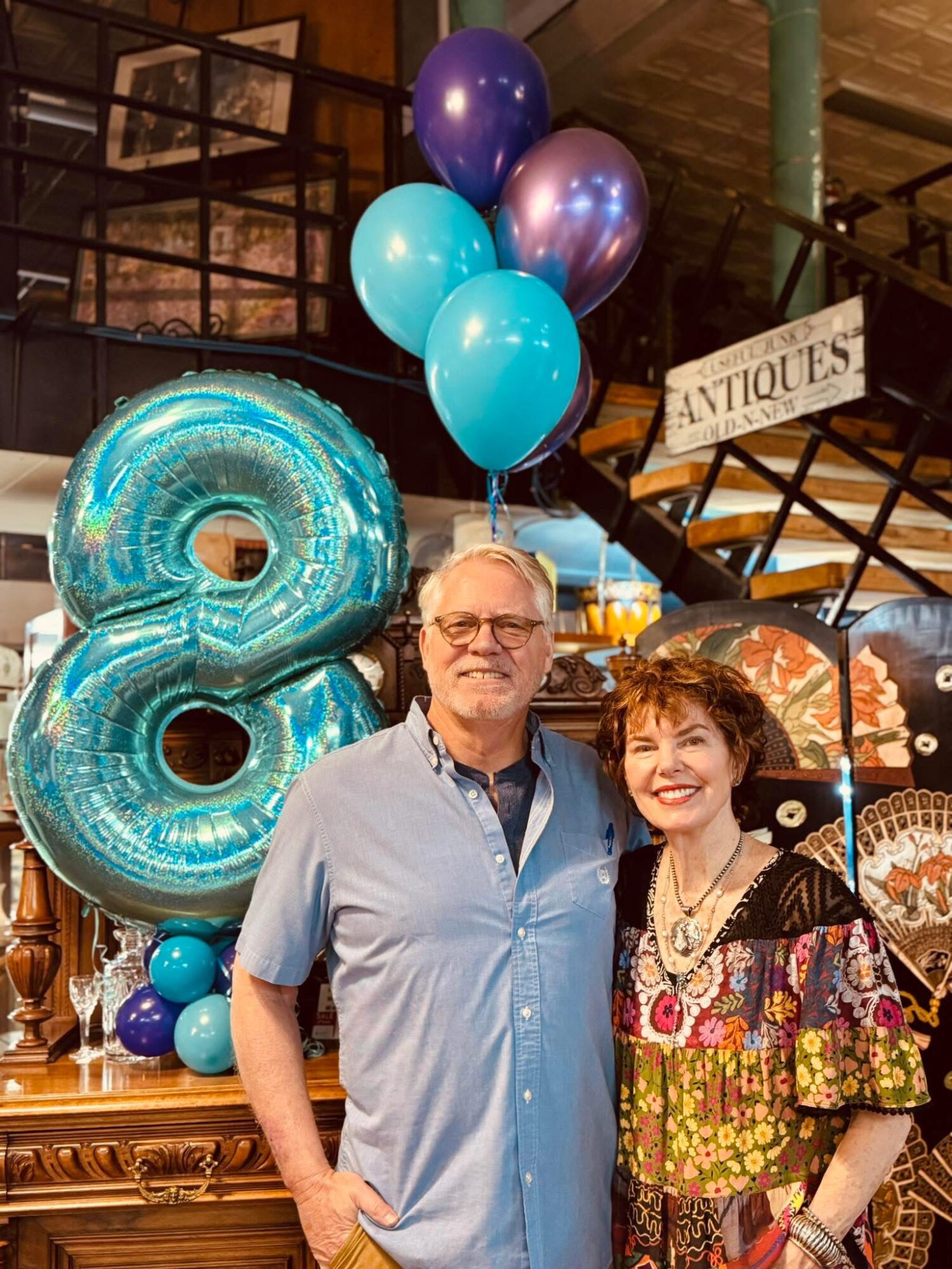 Two people stand in front of balloons and a large number 8 decoration, smiling indoors.