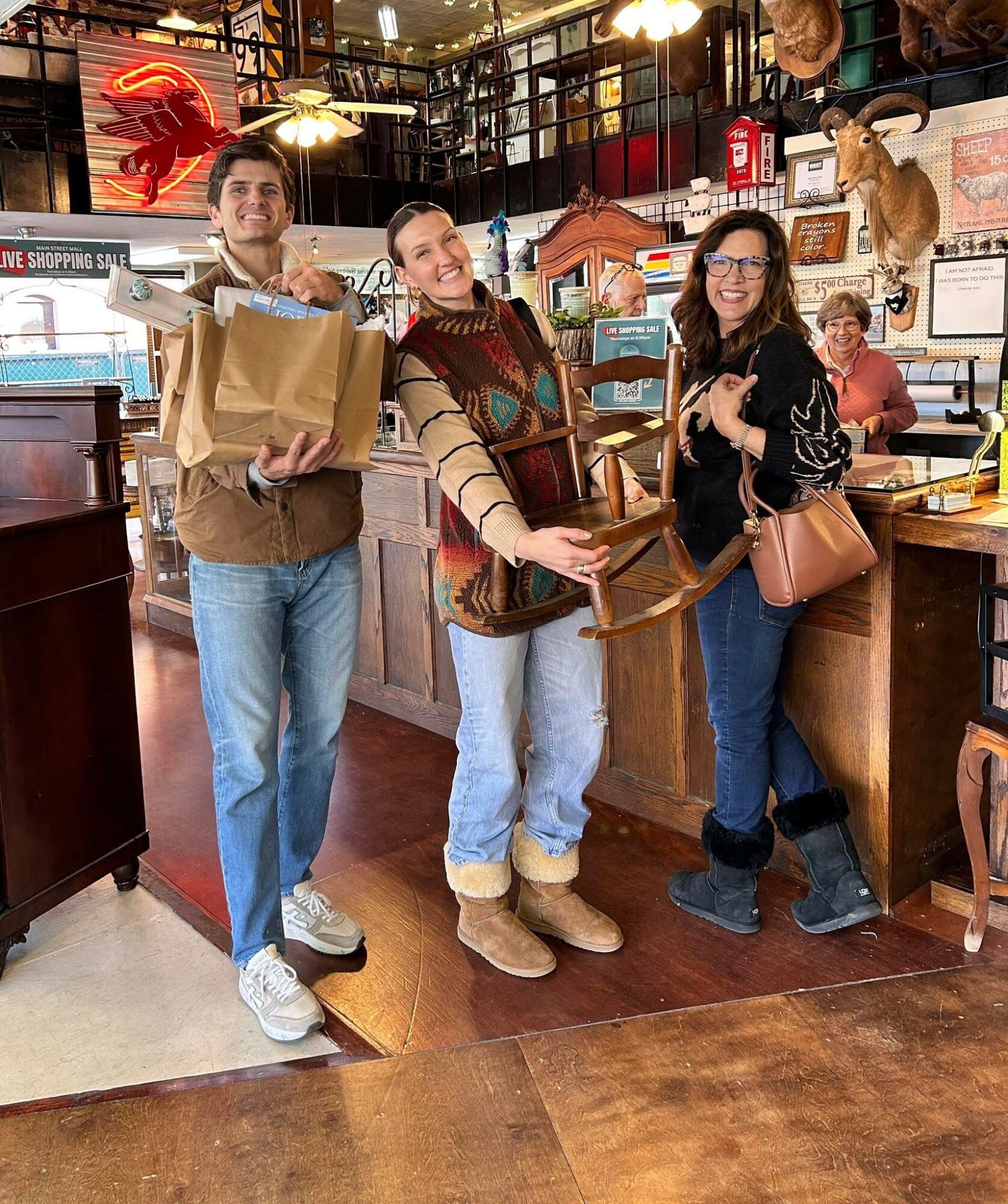 Three people smiling and holding chairs inside a store with various decorations and a person in the background.