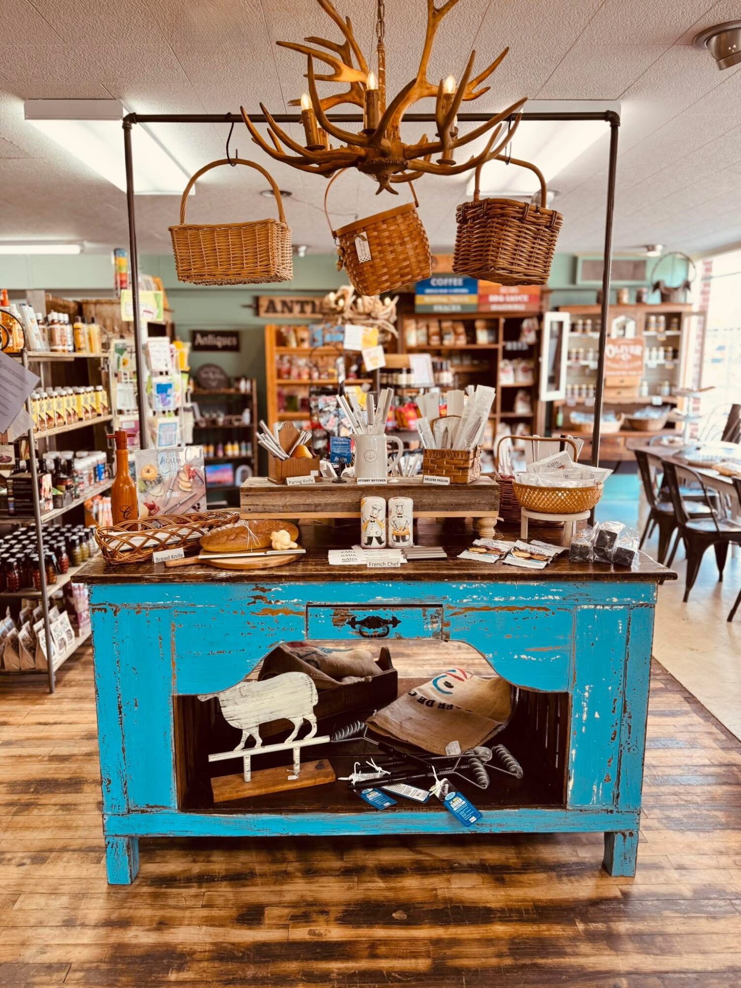 Blue display table with baskets and decorative items inside a store, chandelier with antlers hanging above, wooden floor.