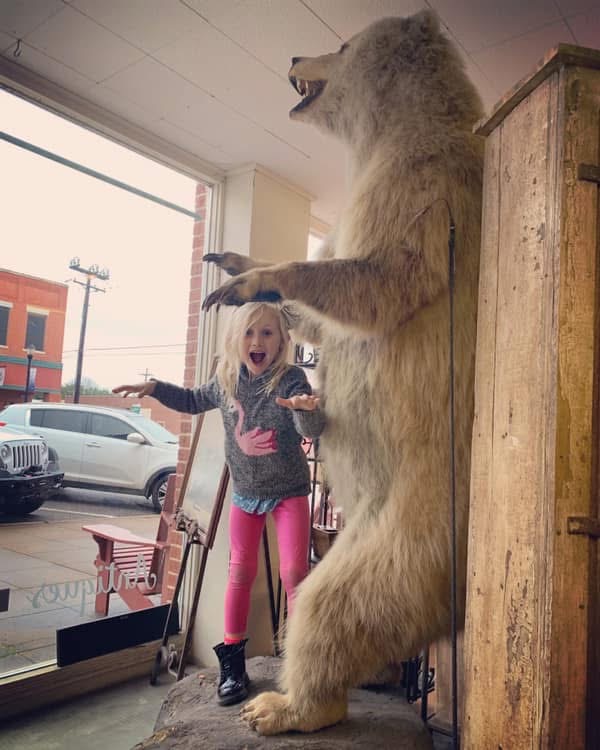 Child with arms raised standing next to a large, standing bear statue inside a building near a window.