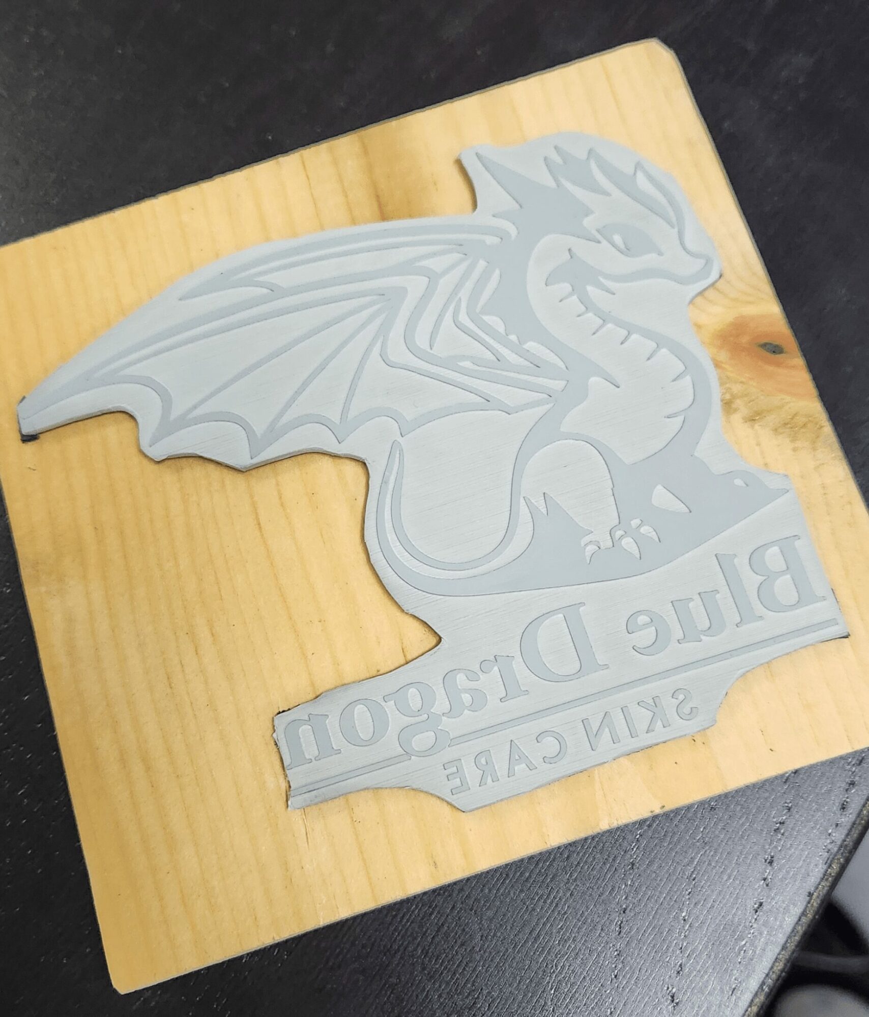 Gray dragon emblem with text on a wooden surface, featuring a dragon with wings and a fierce expression.