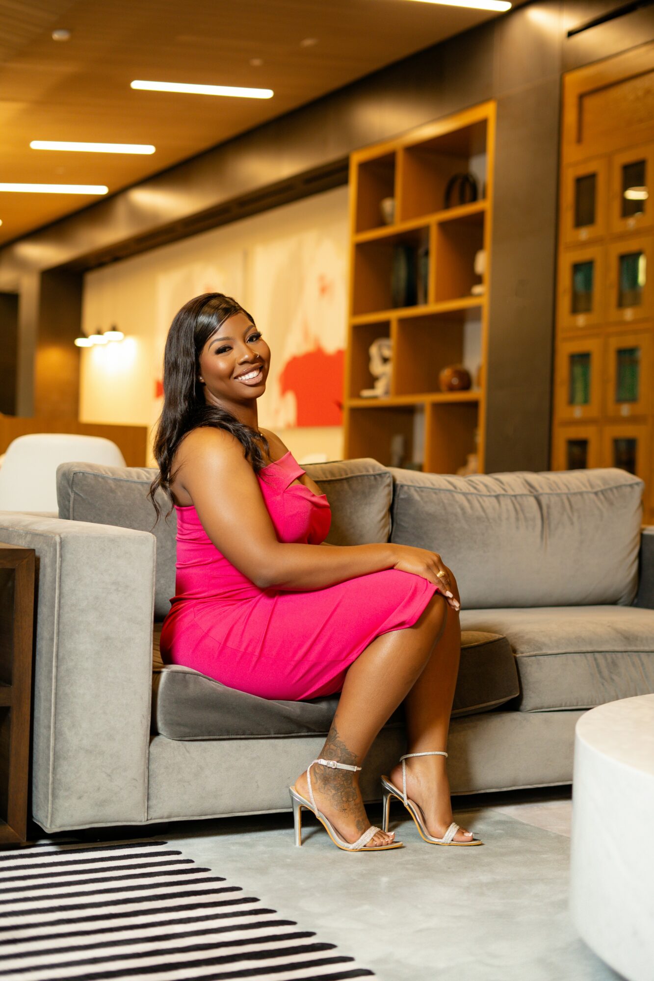 Woman in pink dress sitting on a gray sofa in a modern room with wooden shelves and warm lighting.