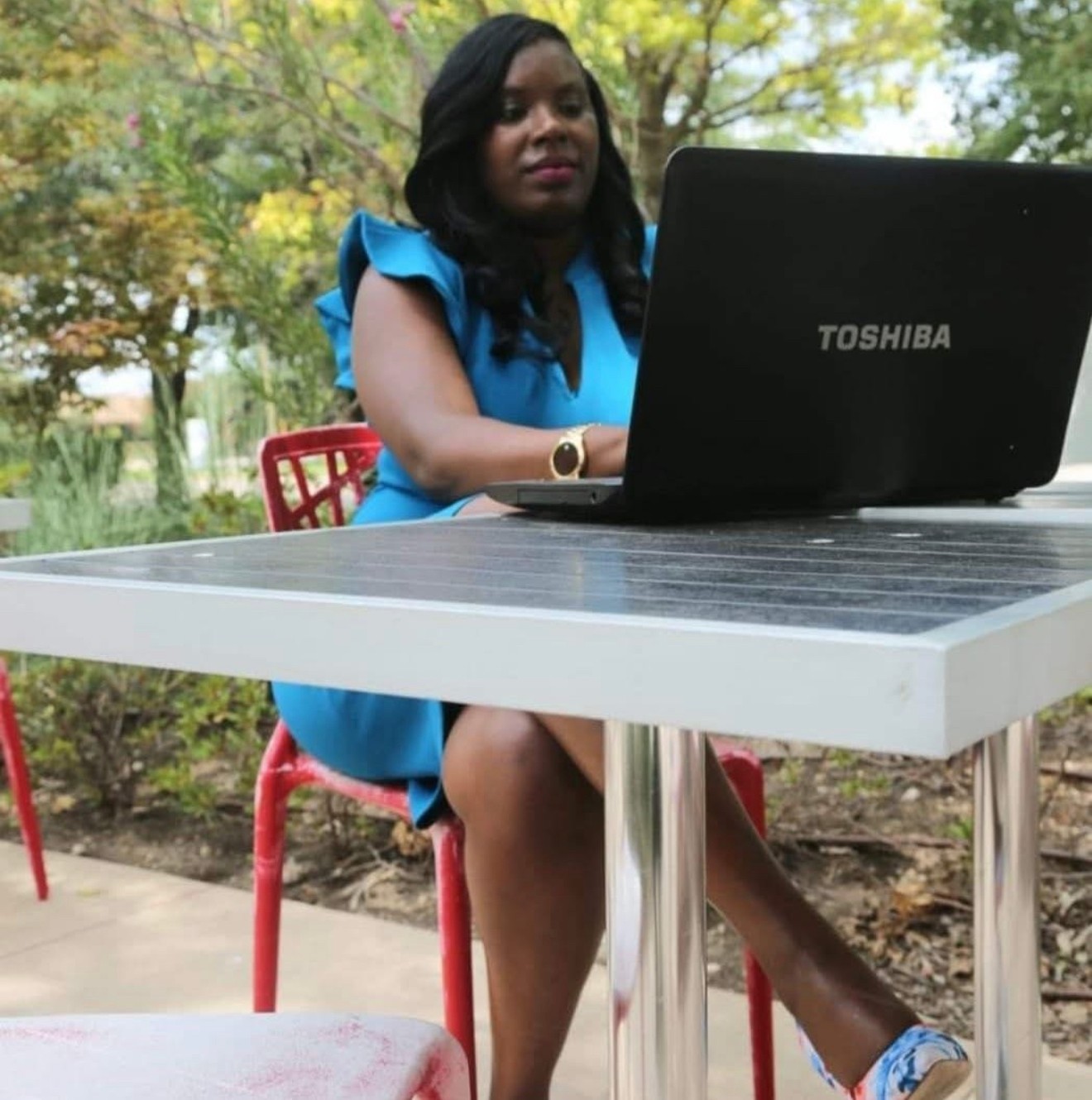 Woman sitting outdoors at a table with a laptop, wearing a blue dress, with trees in the background.