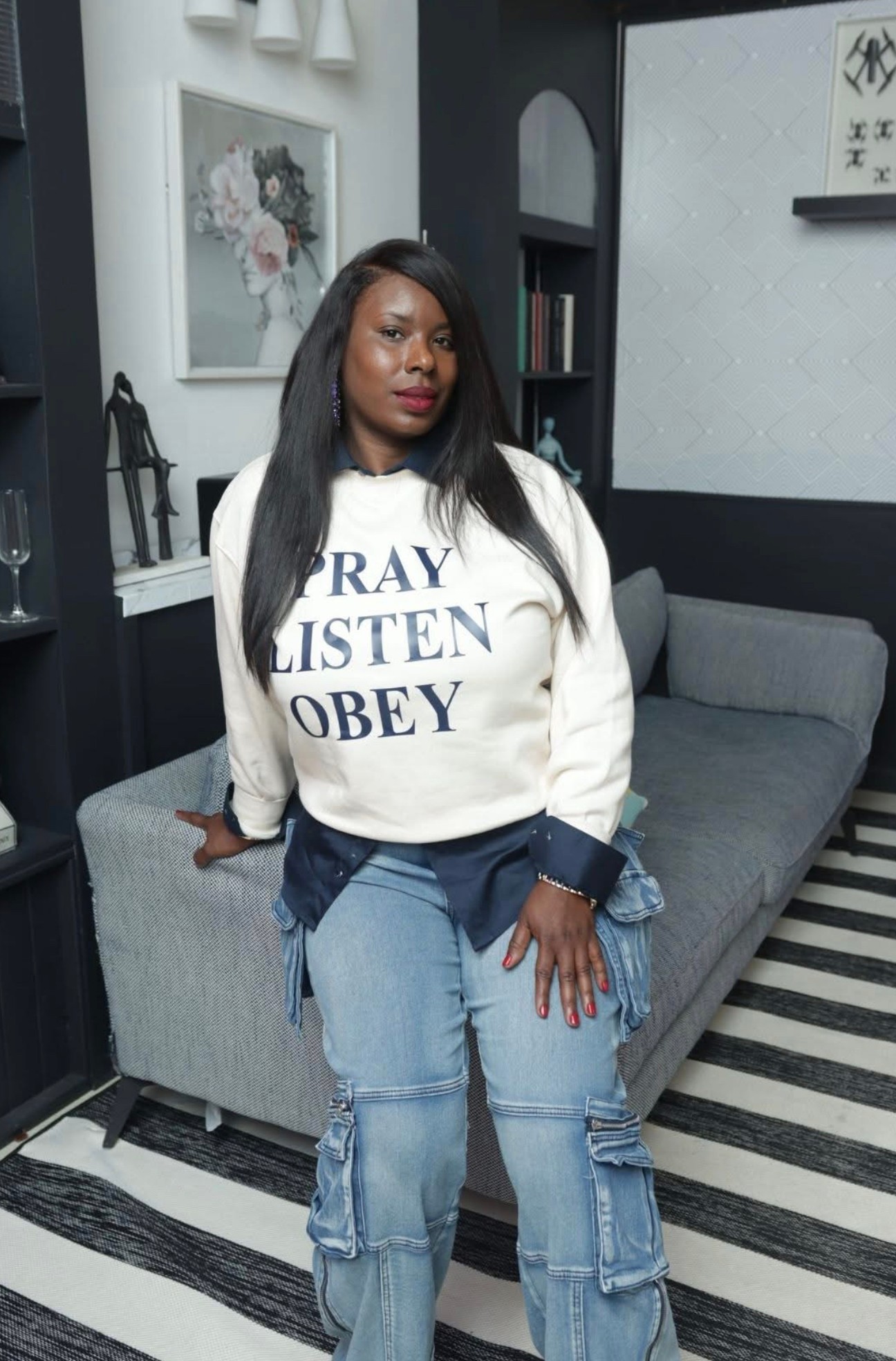 Woman with long black hair wearing a white sweatshirt and jeans, standing in a modern living room.