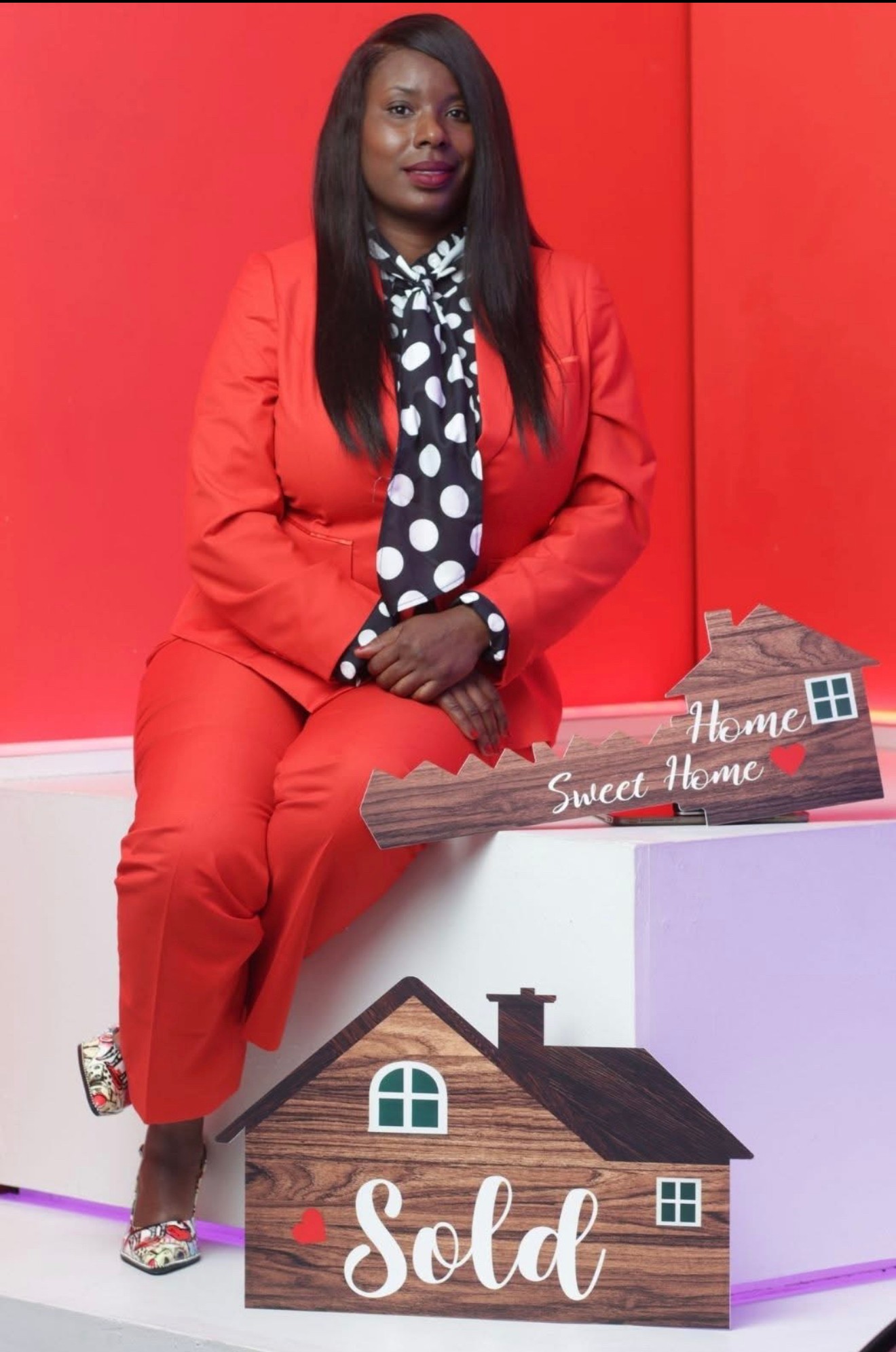 Woman in red suit sitting against red background, holding a house-shaped sign that says 'Sold'.