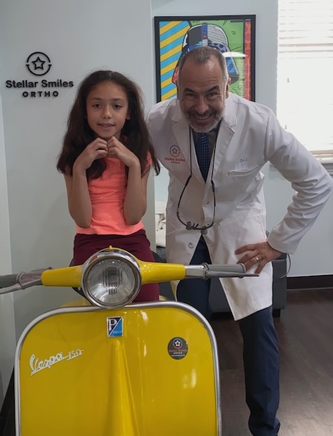 Young girl and male doctor smiling indoors, yellow Vespa scooter in foreground, framed artwork on wall behind.