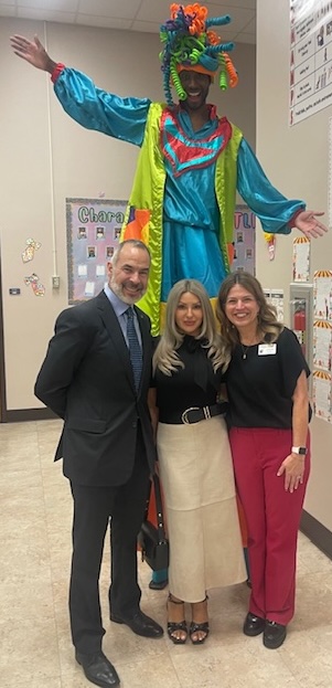 Three people standing in front of a colorful clown statue with arms outstretched in a room with educational posters.