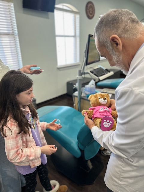 Child and doctor in a medical office with a teddy bear, medical equipment, and a window in the background.