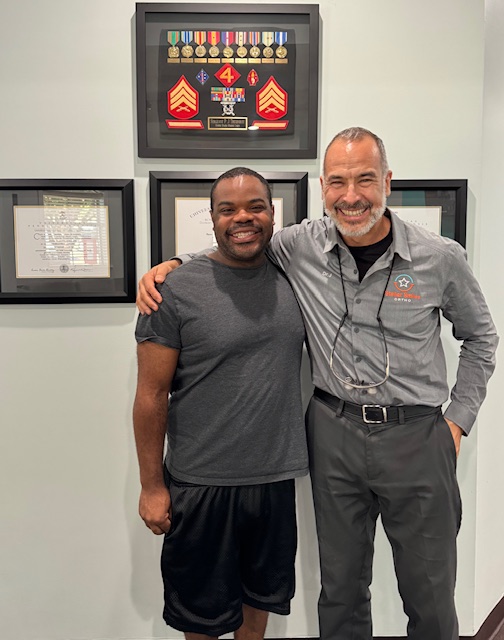Two men standing side by side, smiling, with framed artwork on the wall behind them.