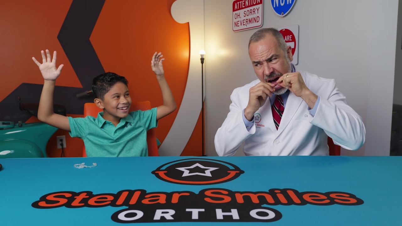 Child and adult sitting at a table with a Stella's Smiles banner, smiling and raising hands, indoor setting.
