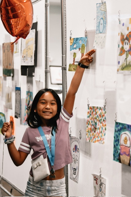 Girl smiling and pointing at artwork on display wall in gallery or classroom setting.