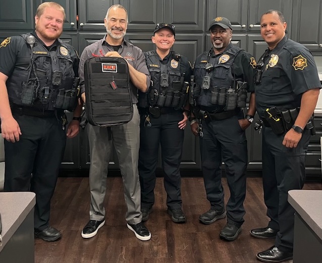 Five police officers and one person holding a backpack standing in front of dark cabinets.