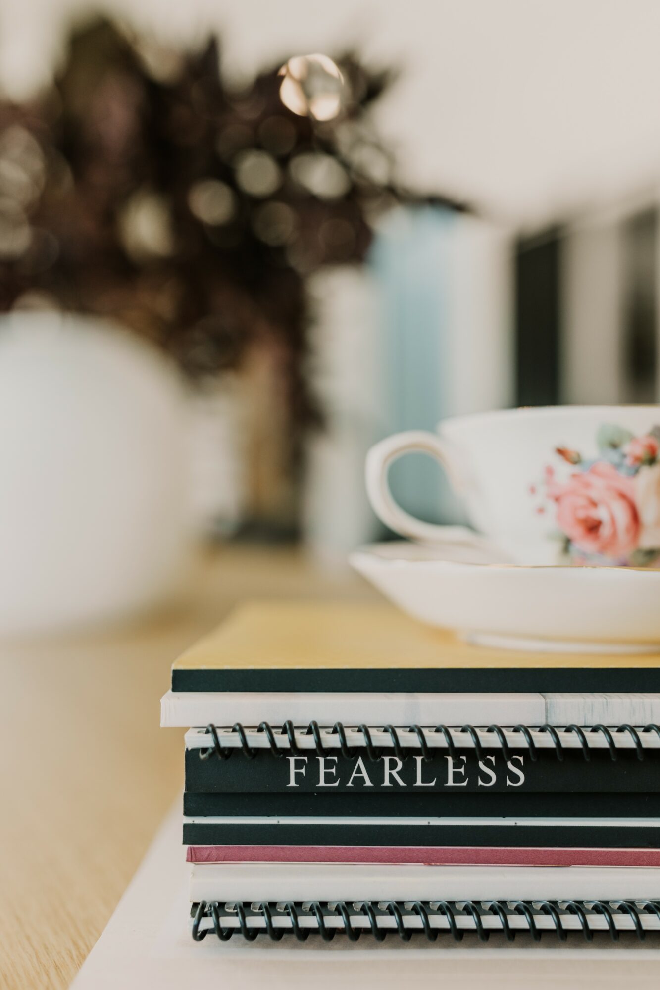 Close-up of a spiral-bound notebook with the word FEARLESS on the cover, placed on a table with a teacup and books in background.