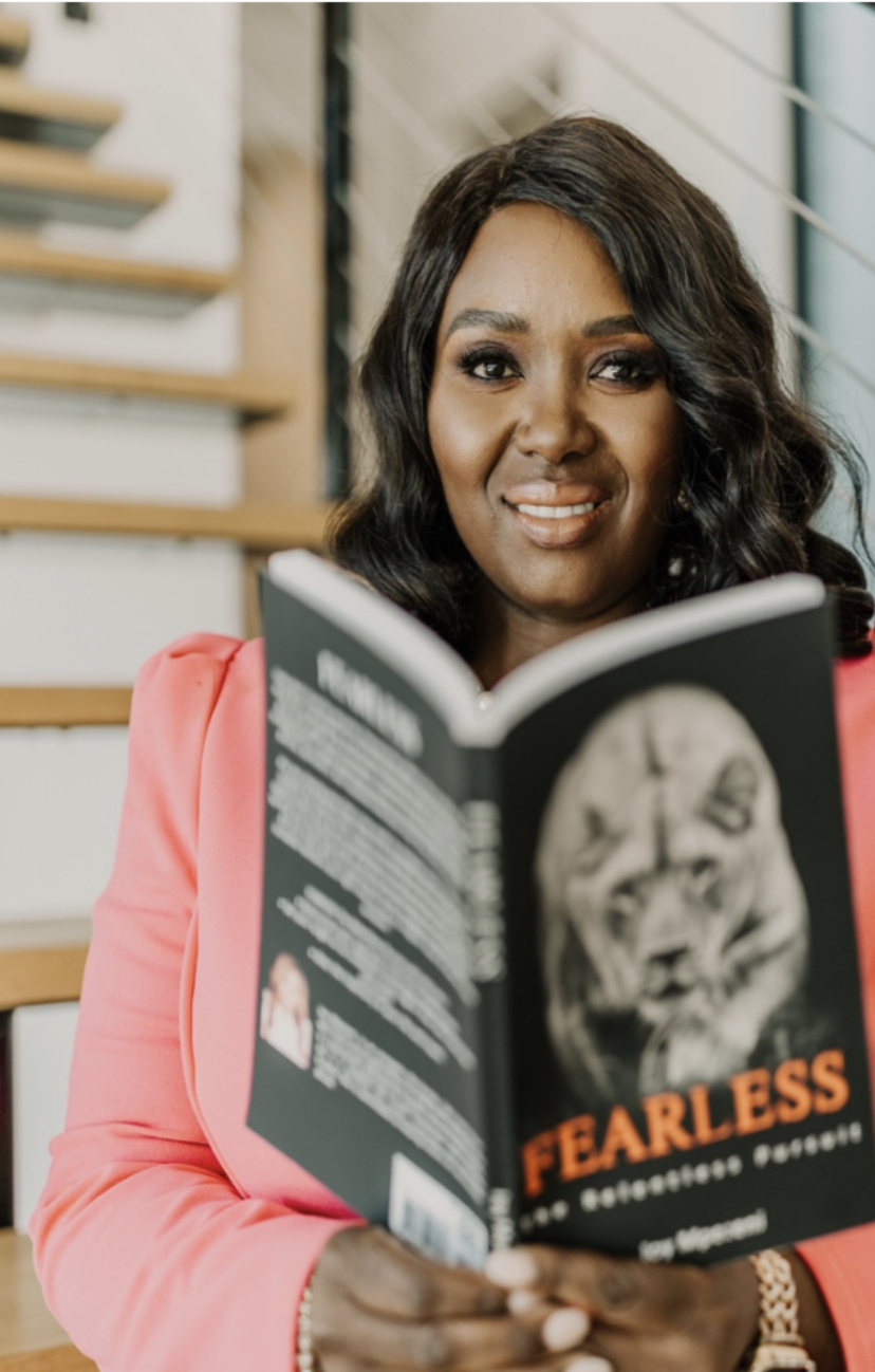 Woman with dark hair in a pink blazer holding a book titled 'Fearless' with a lion illustration, smiling indoors.
