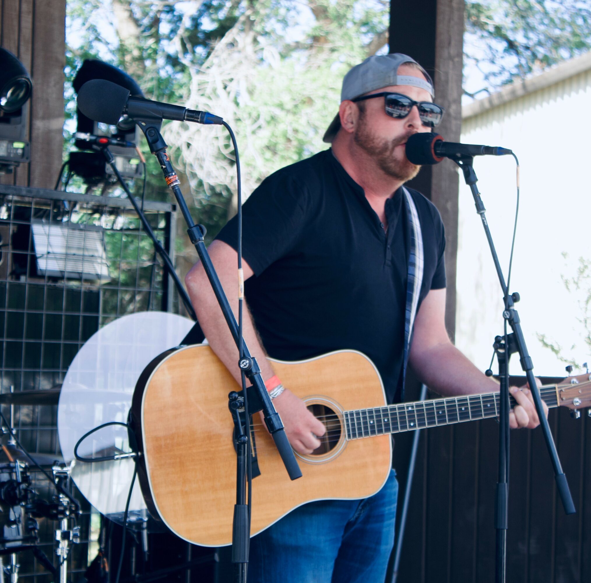 Man wearing sunglasses, cap, and black shirt playing acoustic guitar and singing into microphone outdoors.