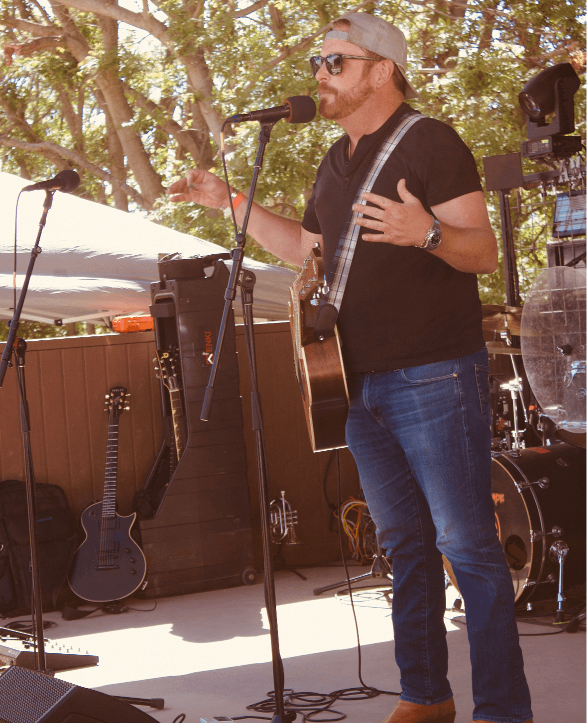Man singing into microphone outdoors, playing guitar, wearing sunglasses, cap, black t-shirt, jeans, surrounded by musical equipment.