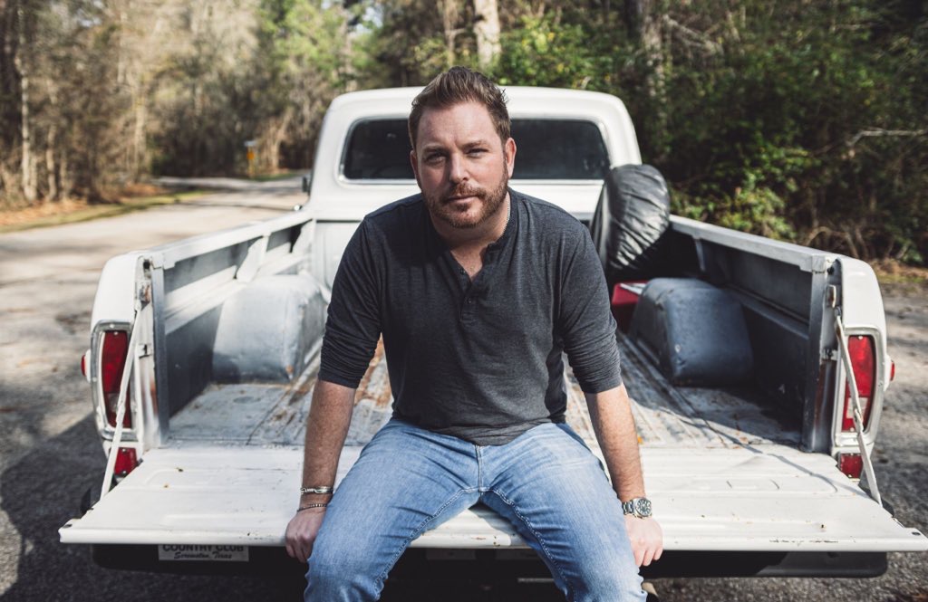 Man sitting on the open tailgate of a white pickup truck outdoors with trees in the background.