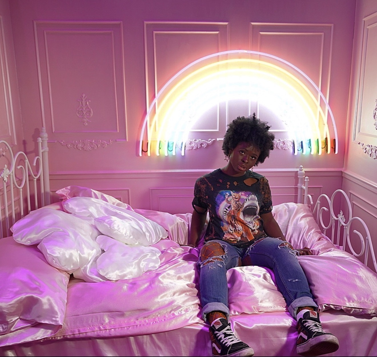 Person sitting on a bed with pink satin sheets and pillows, neon rainbow light on pink wall behind them.