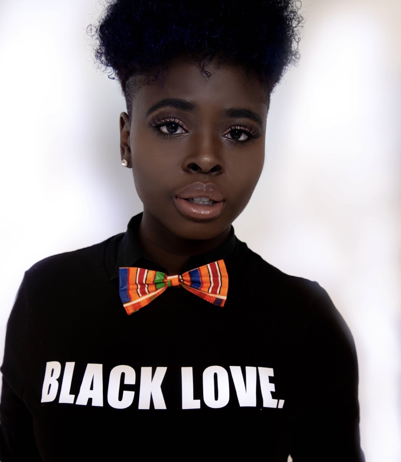 Young woman with natural hair wearing a black shirt with white text and a colorful bow tie.
