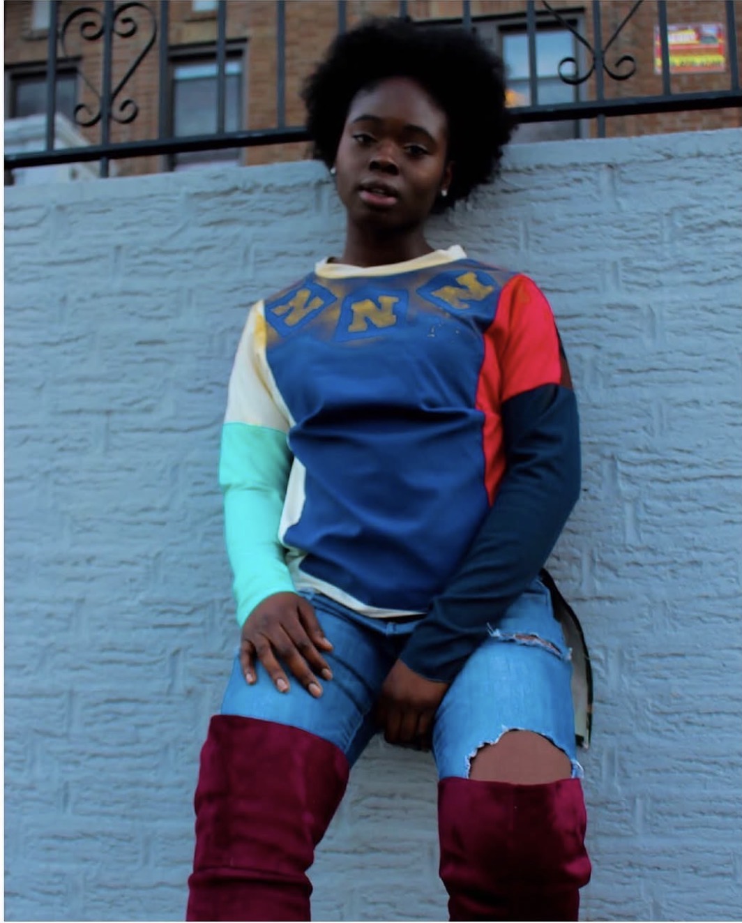 Young person with natural hair standing against a blue wall, wearing a colorful long-sleeve shirt and ripped jeans.