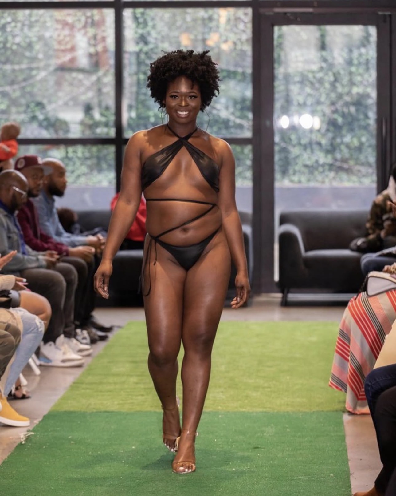 Model walking on runway in black strappy swimsuit, audience seated on both sides, indoor setting with large windows.