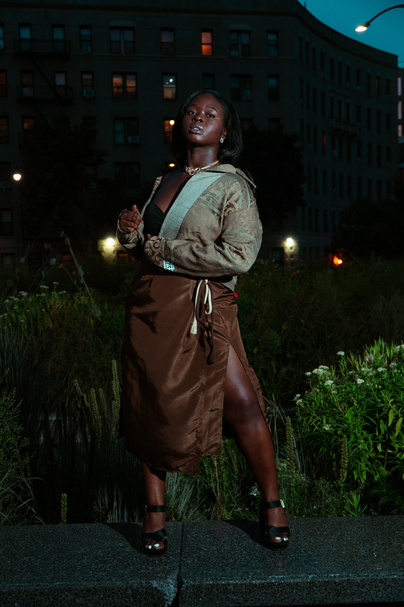 Woman standing outdoors at night in front of a building, wearing a jacket and skirt, with city lights behind her.