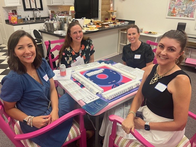 Four women sitting around a table with a cake, smiling, in a room with kitchen and colorful decorations.