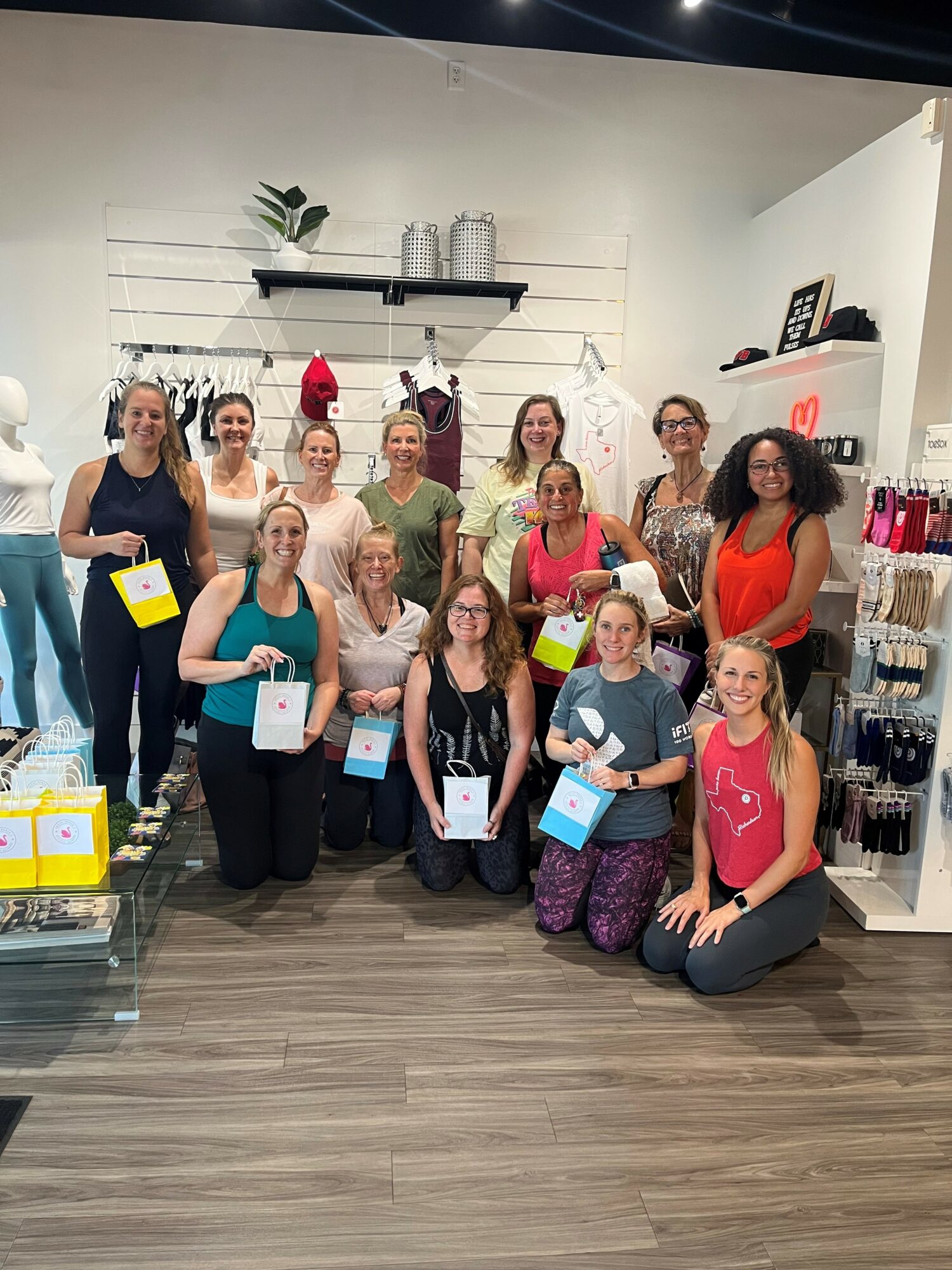 Group of 13 women in a store, some holding shopping bags, smiling, with shelves and products in background.