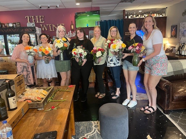 Group of eight women holding bouquets of flowers, standing indoors in a room with bar and TV, smiling.