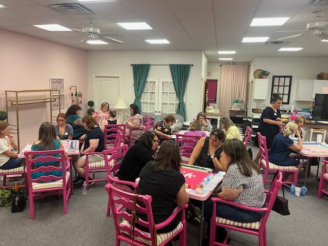 Group of people sitting at tables in a room with pink chairs and green curtains, engaging in activities.