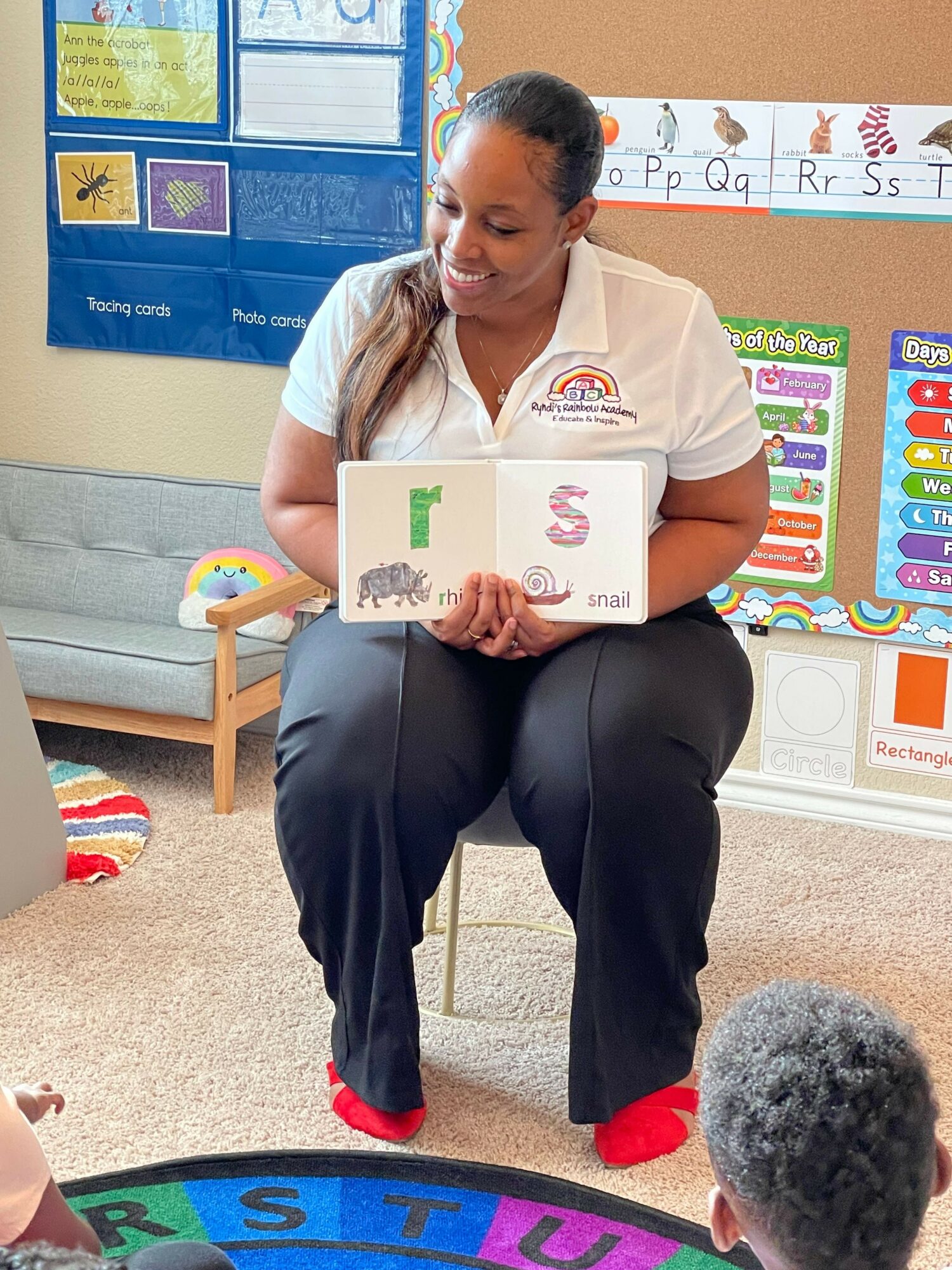Woman sitting on a chair in a classroom, holding a book with animal pictures, with children and educational posters around.