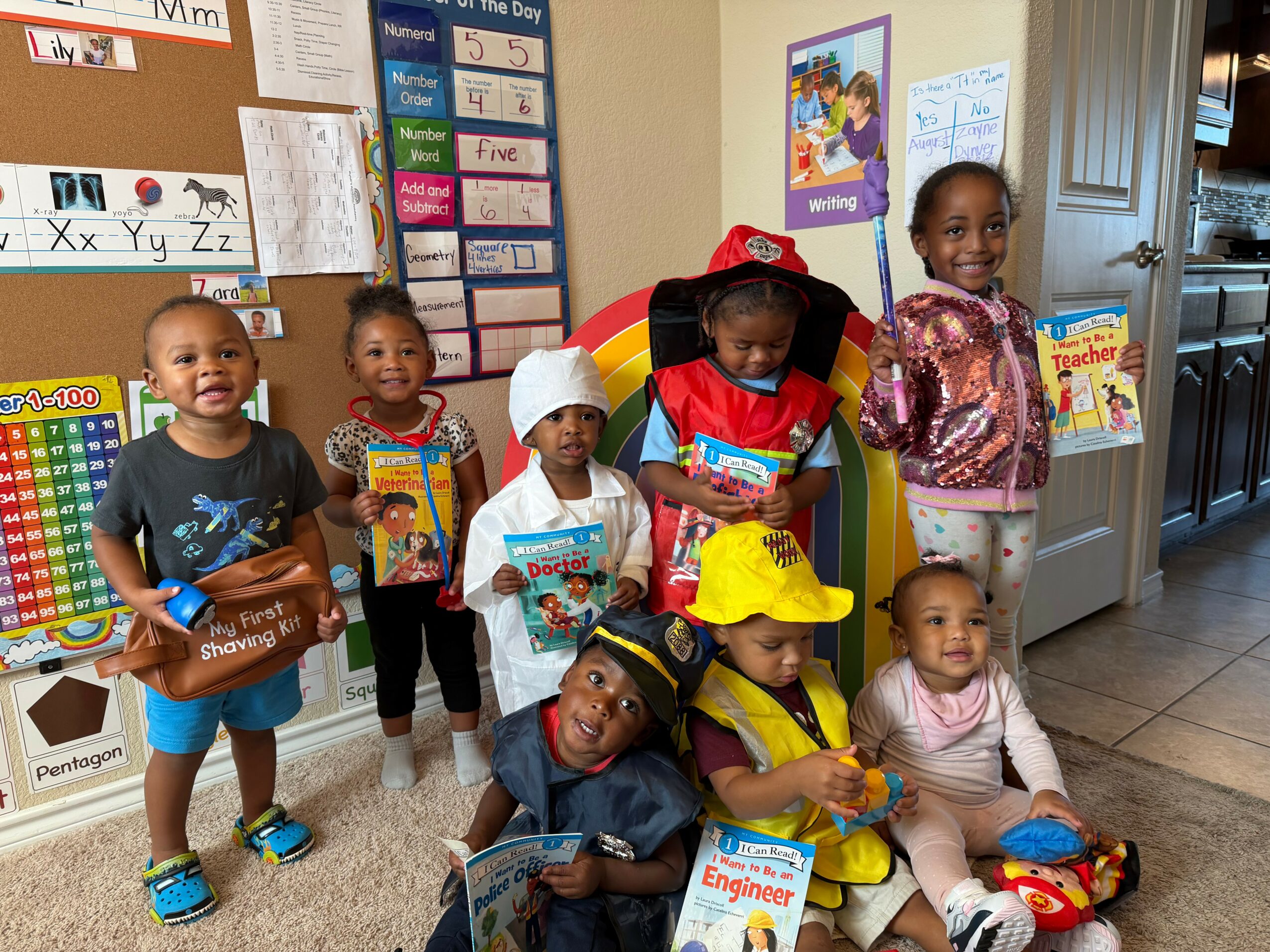Group of seven children in a classroom holding books and toys, some sitting and some standing, with educational posters on wall.