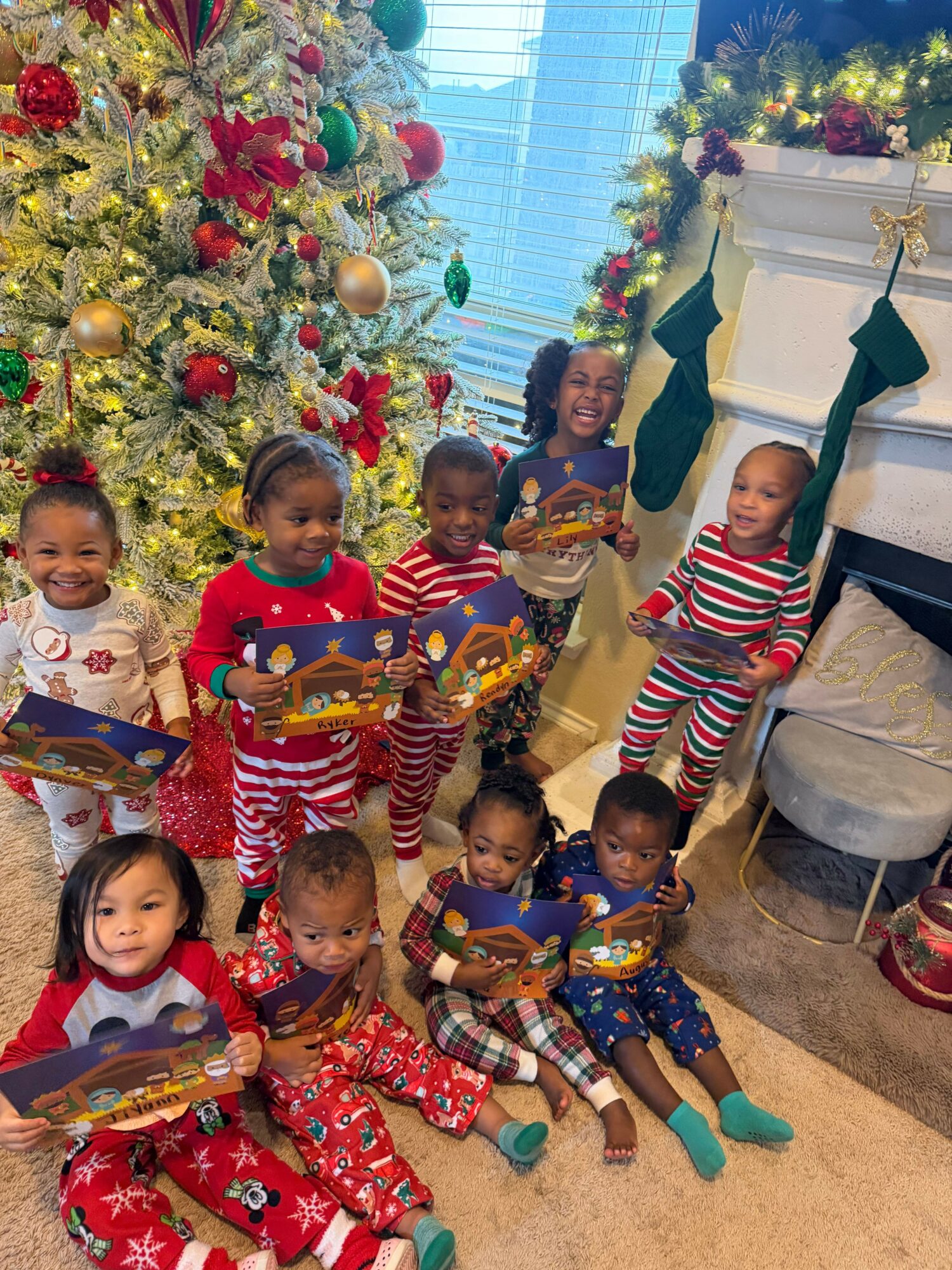 Group of children in Christmas pajamas holding books, standing near decorated Christmas trees and a fireplace.