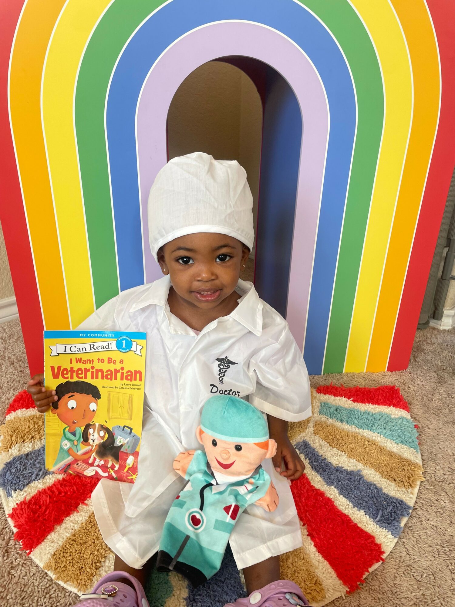 Child dressed as a doctor holding a toy, standing in front of a rainbow backdrop and a colorful rug.