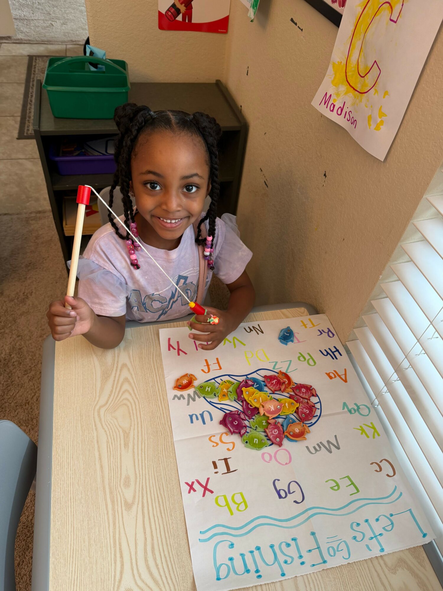 Young girl with braided hair smiling, holding a paintbrush, sitting at a table with a colorful drawing of a heart and flowers.