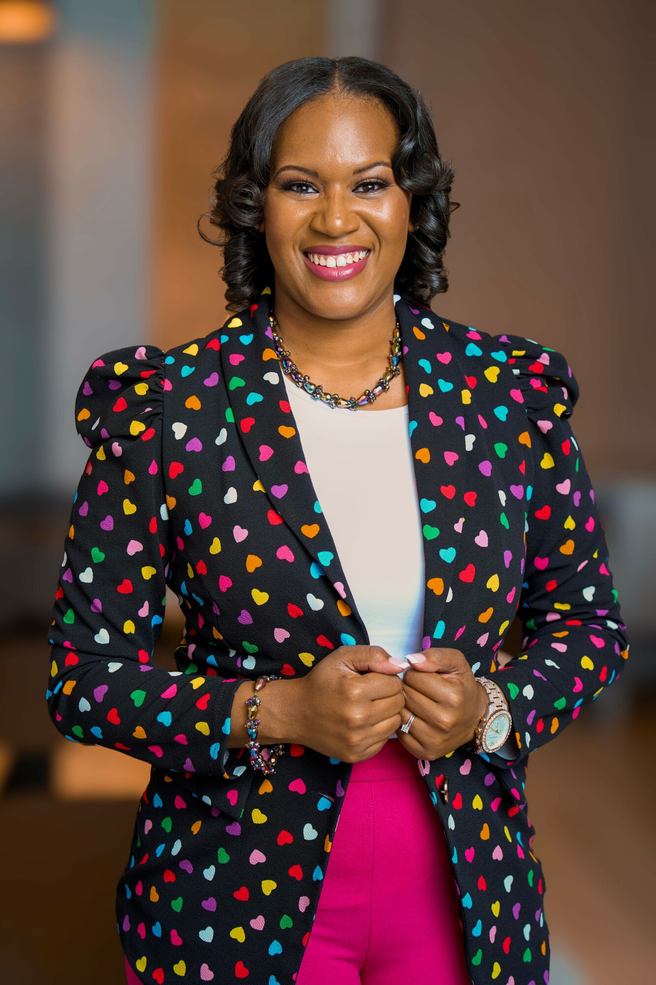 Woman smiling, wearing a colorful heart-patterned blazer and pink pants, holding hands together, indoors.