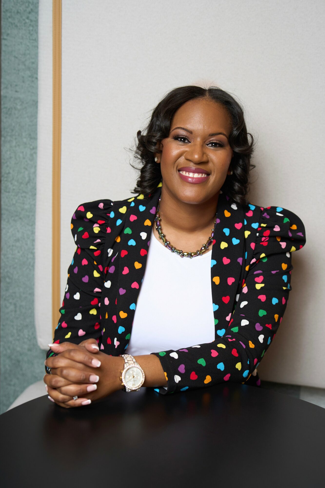 Woman with curly hair smiling, wearing a colorful polka dot blazer and white top, sitting at a table.