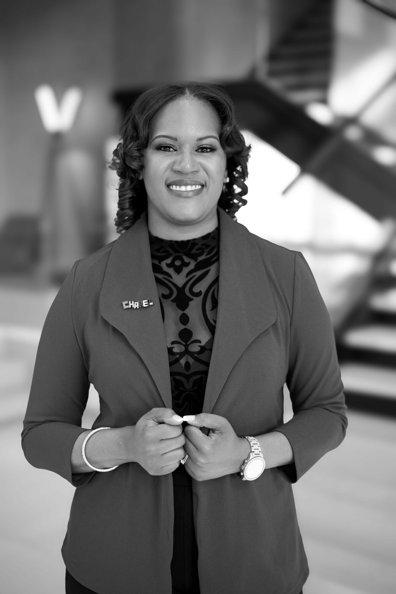 Woman with curly hair in professional attire, smiling, standing indoors with modern architecture background.
