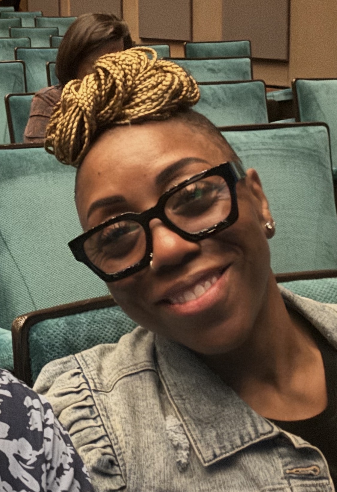 Smiling woman with glasses and a headwrap, sitting in a room with green chairs.