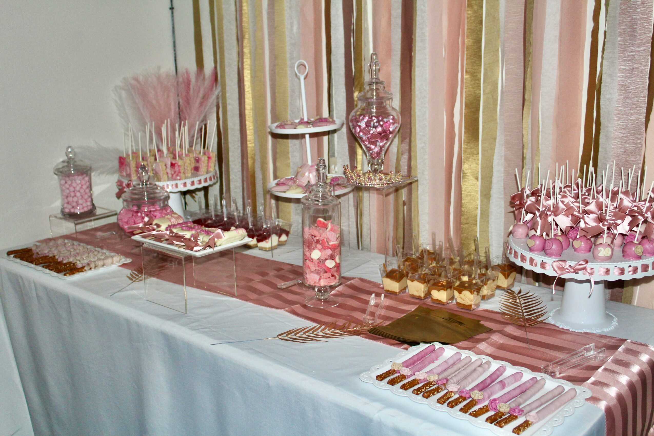 Decorative pink and gold party table with sweets, candies, and treats on a pink striped tablecloth, with pink curtains in background.