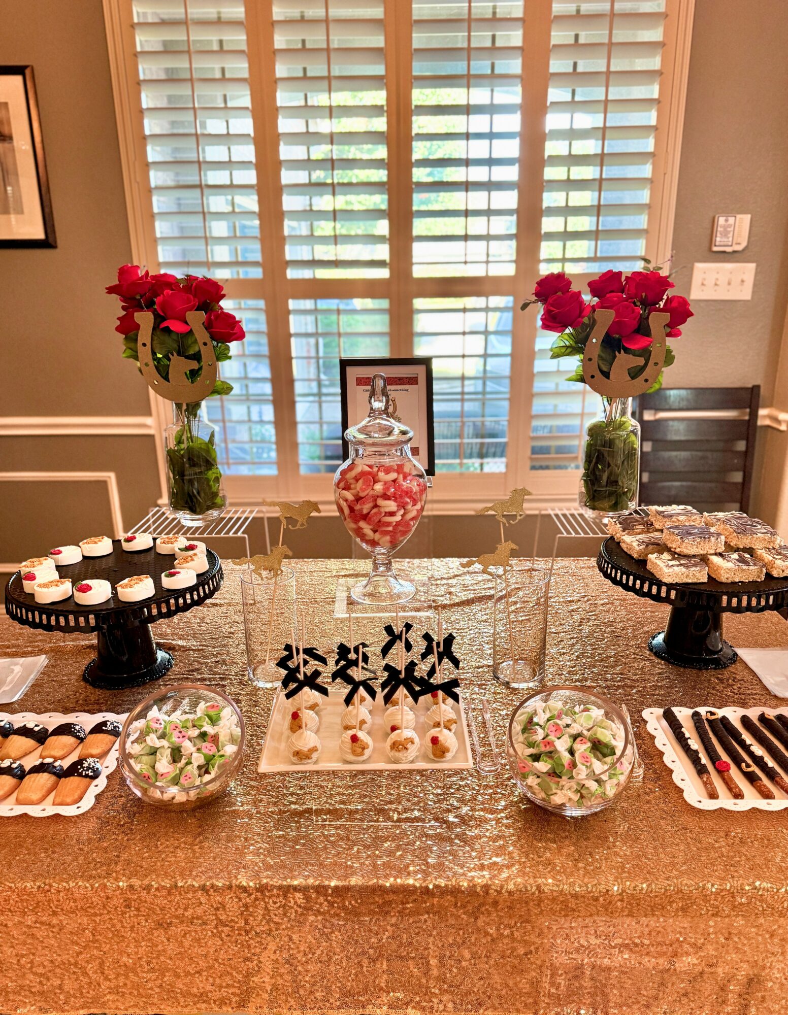 Decorative table with flowers, sweets, and a framed photo in front of window with blinds.