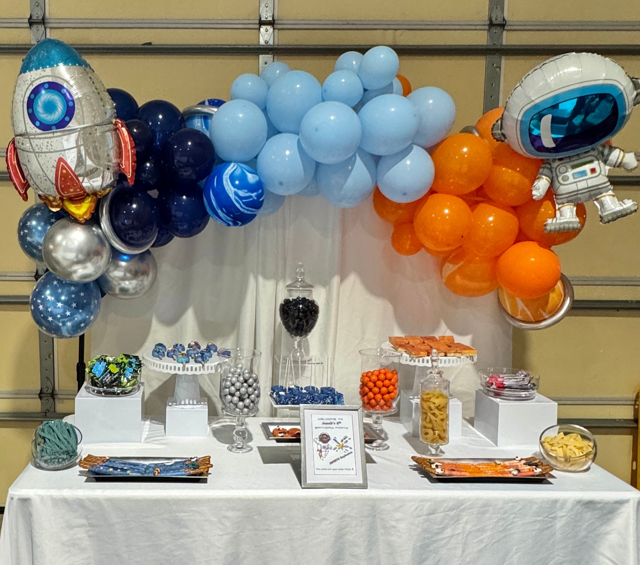 Party table with blue and orange balloons, space-themed decorations, and treats in containers, with a white tablecloth.